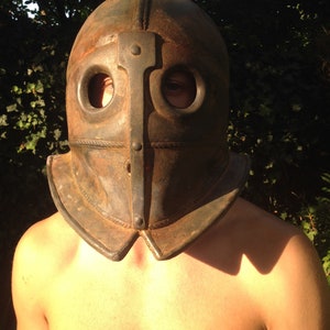 Puede incluir: Un primer plano de una persona que lleva un casco de cuero marr&oacute;n oscuro con dos agujeros para los ojos y una placa de metal en la parte delantera. El casco tiene una parte superior redondeada y una barbilla puntiaguda.
