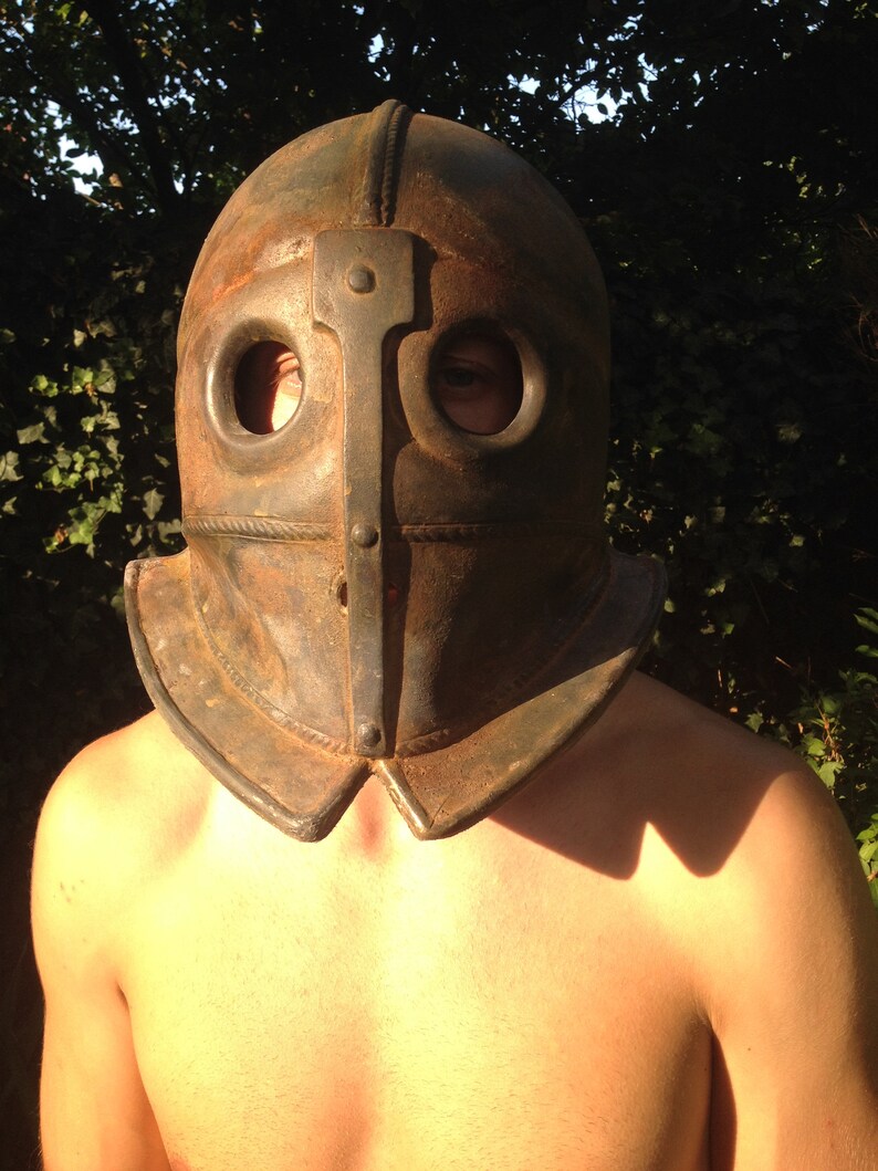 Puede incluir: Un primer plano de una persona que lleva un casco de cuero marr&oacute;n oscuro con dos agujeros para los ojos y una placa de metal en la parte delantera. El casco tiene una parte superior redondeada y una barbilla puntiaguda.