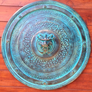 May include: A round, teal-colored metal shield with intricate carvings and a raised, bronze-colored animal head in the center. The shield has a decorative border and is mounted on a wooden surface.