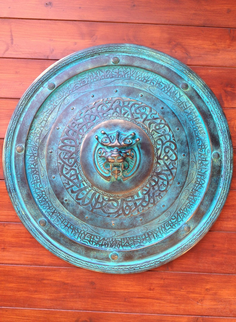 May include: A round, teal-colored metal shield with intricate carvings and a raised, bronze-colored animal head in the center. The shield has a decorative border and is mounted on a wooden surface.