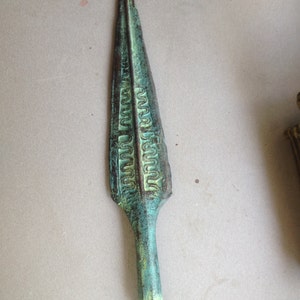 Peut inclure: Une pointe de lance en bronze vert avec des motifs complexes gravés à la surface. La pointe de lance est pointue en haut et a une base arrondie.