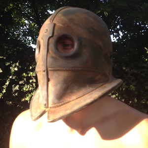 Puede incluir: Un casco de cuero marr&oacute;n con detalles met&aacute;licos y dos agujeros para los ojos. El casco lo lleva una persona con los hombros descubiertos.