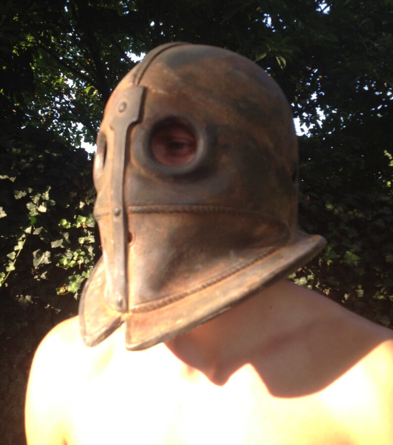 Puede incluir: Un casco de cuero marr&oacute;n con detalles met&aacute;licos y dos agujeros para los ojos. El casco lo lleva una persona con los hombros descubiertos.
