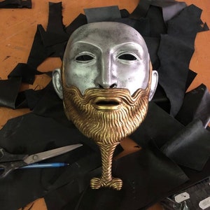 May include: A silver and gold mask with a beard and a long neck. The mask is on a black surface.