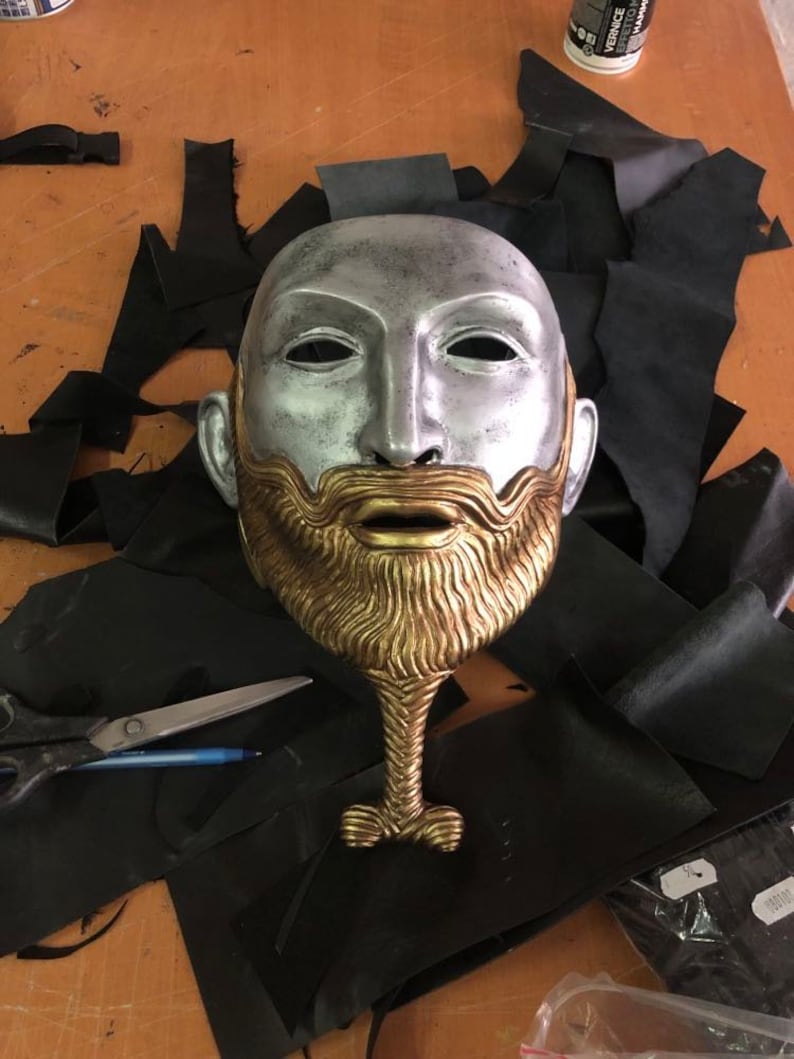 May include: A silver and gold mask with a beard and a long neck. The mask is on a black surface.