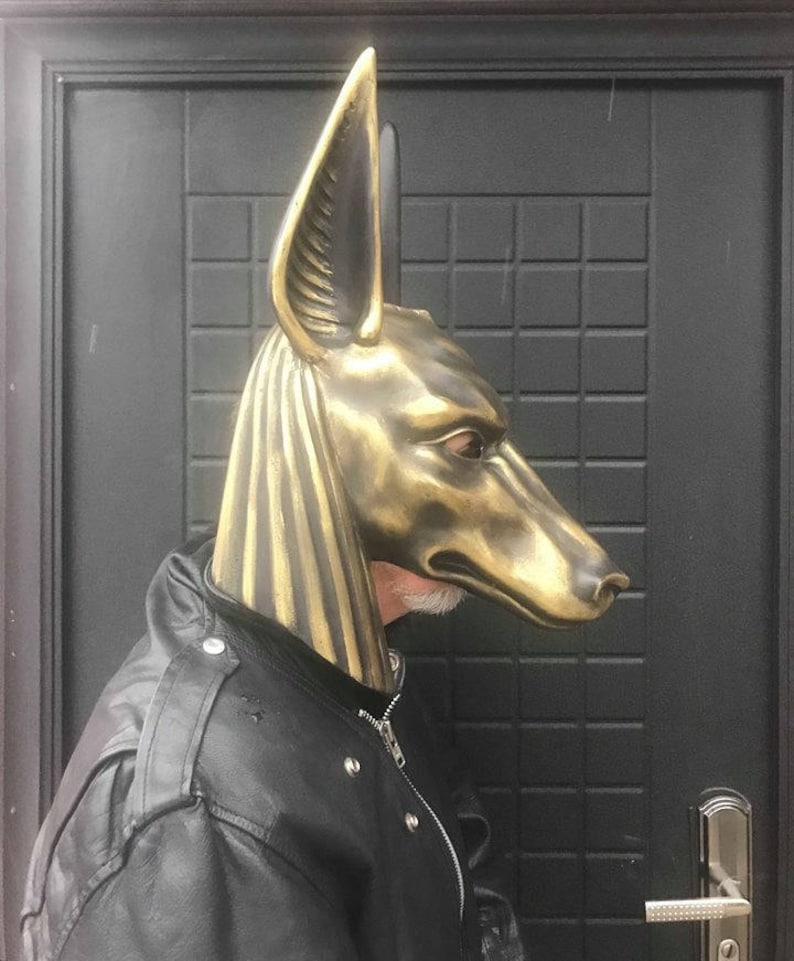 Puede incluir: Una m&aacute;scara de Anubis de color dorado con detalles precisos, usada por una persona con una chaqueta de cuero negra.