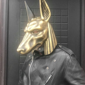 Puede incluir: Una m&aacute;scara de Anubis de color dorado con un dise&ntilde;o detallado. La m&aacute;scara la lleva puesta una persona que lleva una chaqueta de cuero negra.