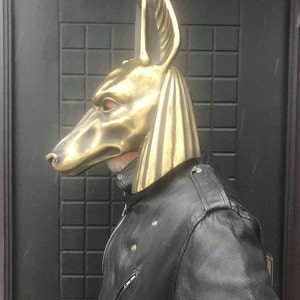 Puede incluir: Una m&aacute;scara de Anubis de color dorado con un dise&ntilde;o detallado. La m&aacute;scara la lleva puesta una persona que lleva una chaqueta de cuero negra.