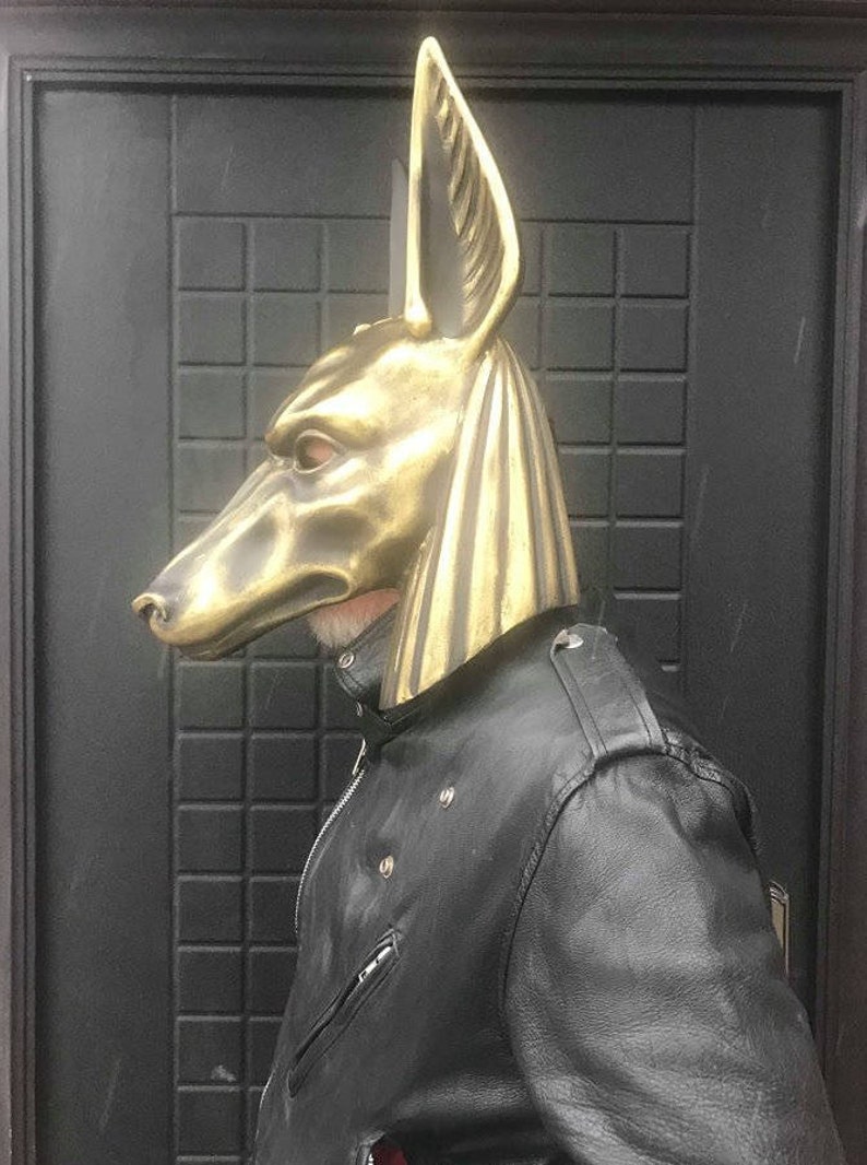 Puede incluir: Una m&aacute;scara de Anubis de color dorado con un dise&ntilde;o detallado. La m&aacute;scara la lleva puesta una persona que lleva una chaqueta de cuero negra.