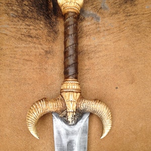 May include: A silver sword with a brown leather wrapped handle and a decorative ivory-colored pommel and guard. The guard is shaped like two ram horns.