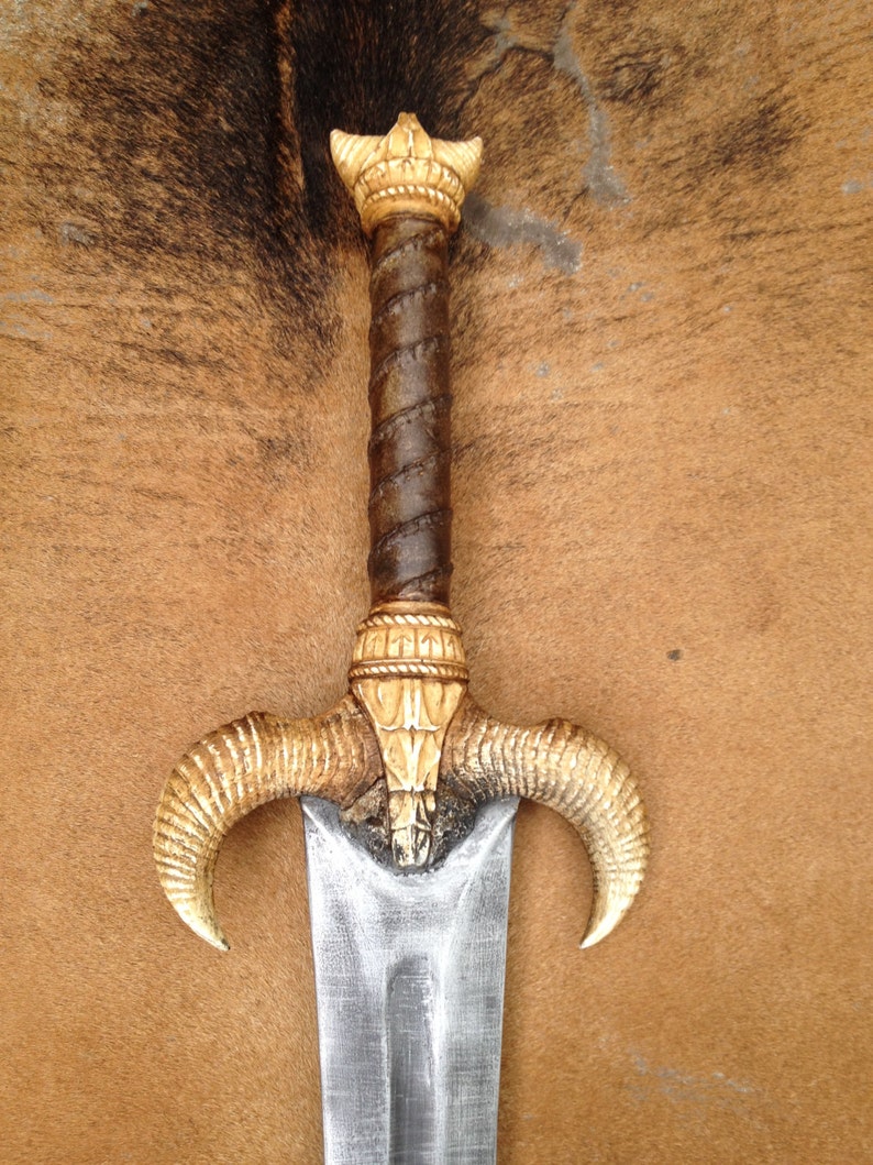 May include: A silver sword with a brown leather wrapped handle and a decorative ivory-colored pommel and guard. The guard is shaped like two ram horns.