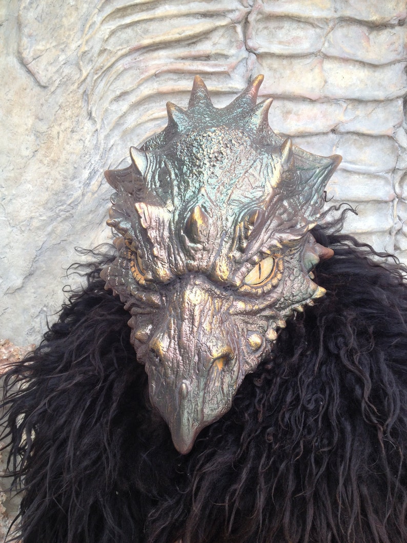 K&ouml;nnte beinhalten: Eine detaillierte, realistische Drachenmaske mit einer strukturierten, metallischen Oberfl&auml;che. Die Maske hat eine lange Schnauze, gro&szlig;e Augen und eine schuppige Textur. Die Maske wird &uuml;ber einem schwarzen, pelzigen Kleidungsst&uuml;ck getragen.