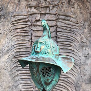 May include: A green metal gladiator helmet with a face mask and a plume on top. The helmet is displayed in front of a textured wall.