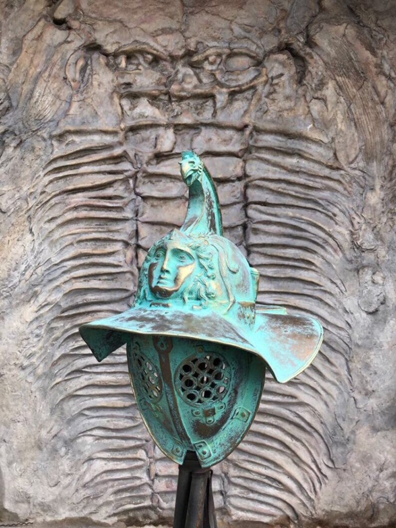 May include: A green metal gladiator helmet with a face mask and a plume on top. The helmet is displayed in front of a textured wall.