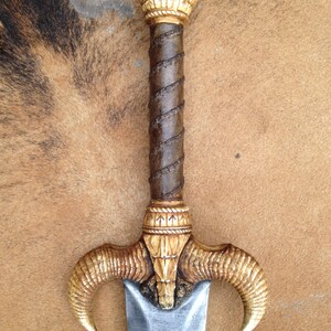 May include: A decorative sword with a gold and brown handle and a silver blade. The handle is adorned with intricate carvings and the blade is sharp and pointed.