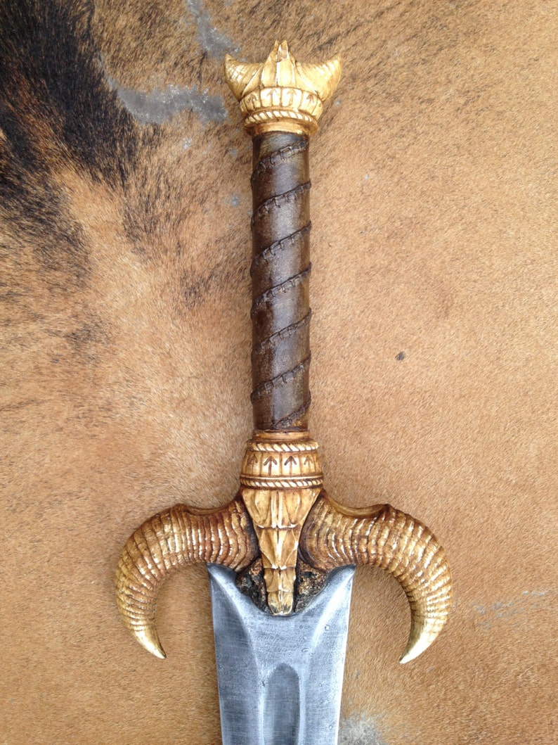 May include: A decorative sword with a gold and brown handle and a silver blade. The handle is adorned with intricate carvings and the blade is sharp and pointed.