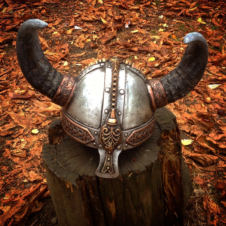 May include: A silver Viking helmet with horns and intricate brass detailing, resting on a wooden stump surrounded by fallen leaves.
