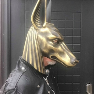 Puede incluir: Una m&aacute;scara de Anubis de color dorado con detalles elaborados, usada por una persona con una chaqueta de cuero negra.
