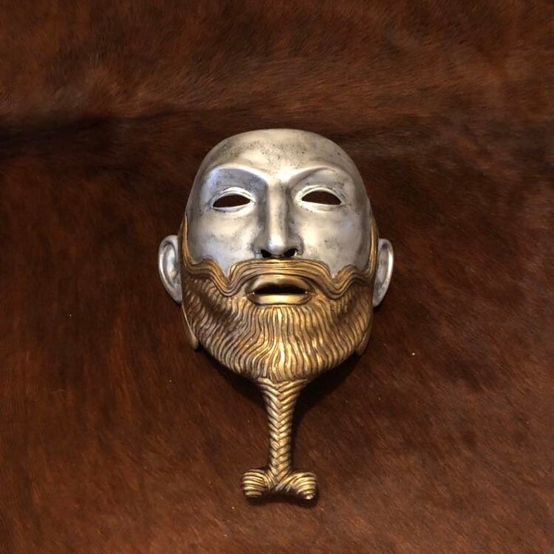 May include: A silver and gold mask with a beard and a long, braided beard extension. The mask has a human face with a serious expression.