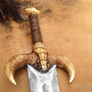 May include: A decorative sword with a gold-colored handle and a silver blade. The handle is shaped like a ram's head with two curved horns.