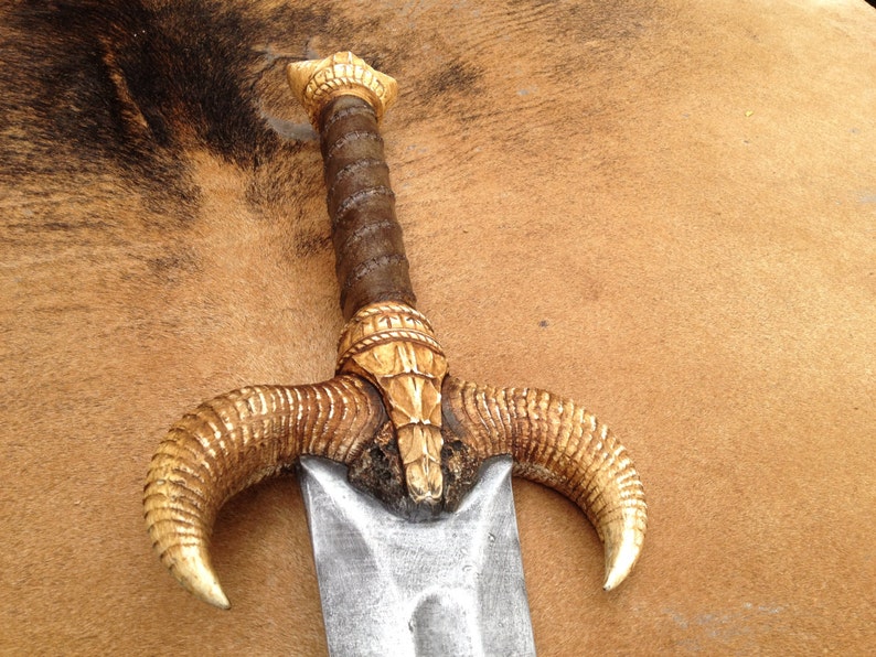 May include: A decorative sword with a gold-colored handle and a silver blade. The handle is shaped like a ram's head with two curved horns.