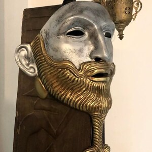 May include: A silver and gold mask with a beard and a detailed design. The mask is mounted on a wooden background.