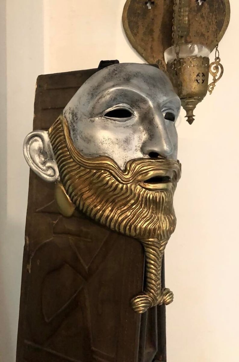 May include: A silver and gold mask with a beard and a detailed design. The mask is mounted on a wooden background.