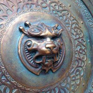 May include: A circular metal shield with an intricate design. The shield is decorated with a raised, detailed image of a lion's head in the center. The lion's head is made of a bronze-colored metal and has a textured surface.