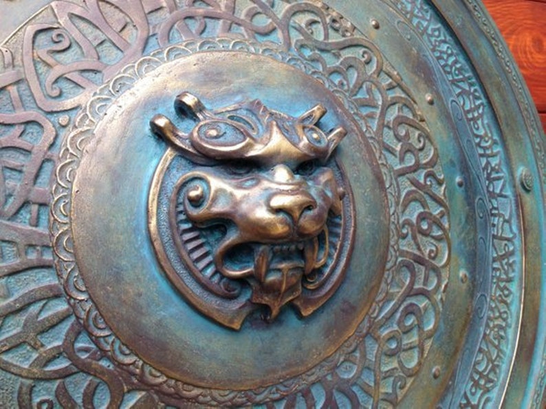 May include: A circular metal shield with an intricate design. The shield is decorated with a raised, detailed image of a lion's head in the center. The lion's head is made of a bronze-colored metal and has a textured surface.
