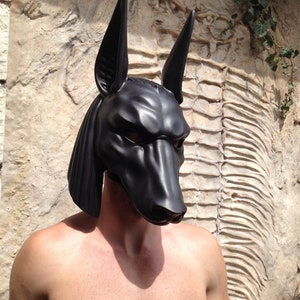 May include: A black, realistic-looking Anubis mask with a textured surface. The mask is worn by a person with bare shoulders and chest.