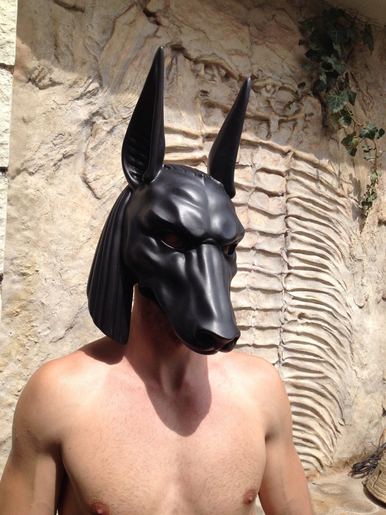 May include: A black, realistic-looking Anubis mask with a textured surface. The mask is worn by a person with bare shoulders and chest.