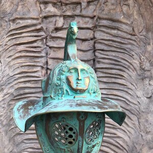 May include: A green bronze gladiator helmet with a face sculpted on the top. The helmet has a large brim and a face mask with a grid pattern.