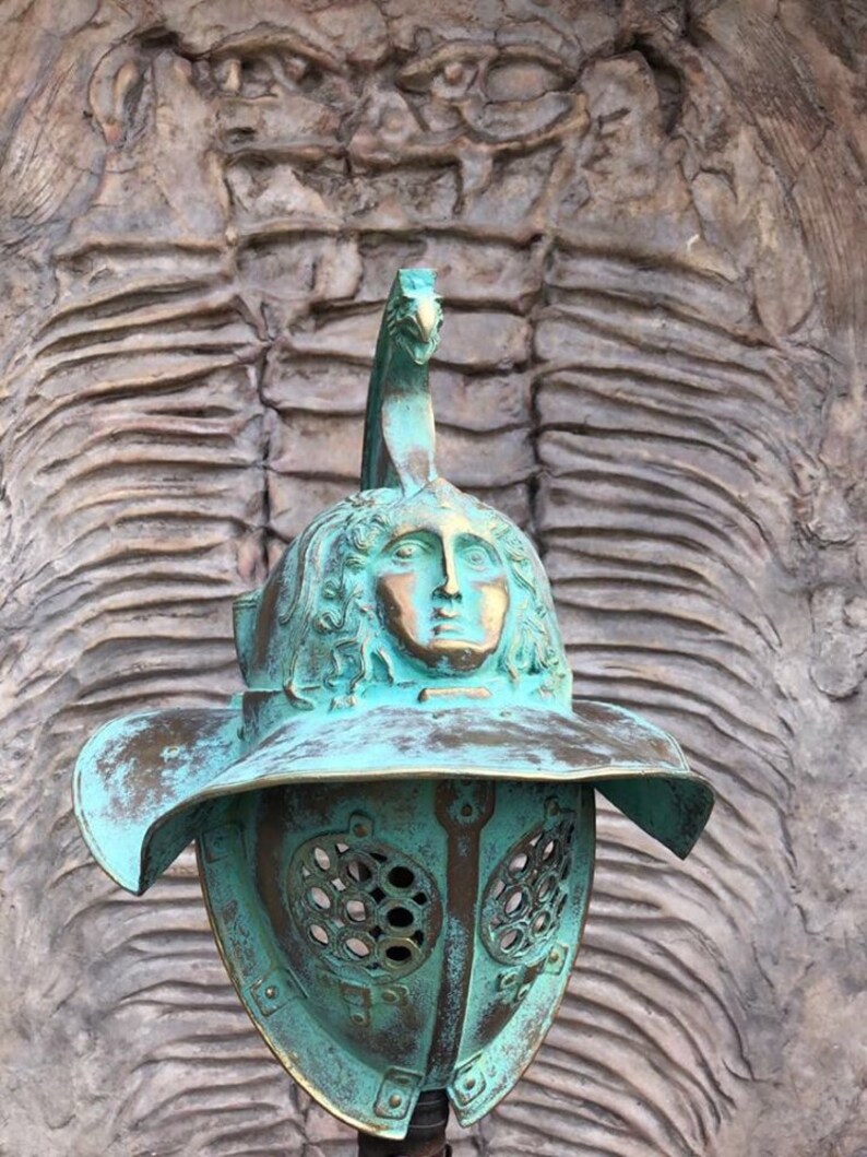 May include: A green bronze gladiator helmet with a face sculpted on the top. The helmet has a large brim and a face mask with a grid pattern.