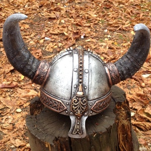 May include: A silver and copper Viking helmet with horns. The helmet has intricate details and a decorative design.