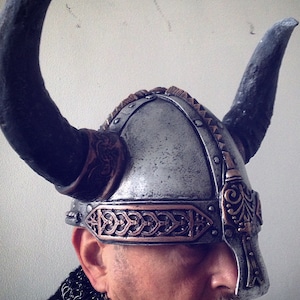 May include: A silver and copper Viking helmet with horns, worn by a person with a beard. The helmet has intricate details and a braided band.