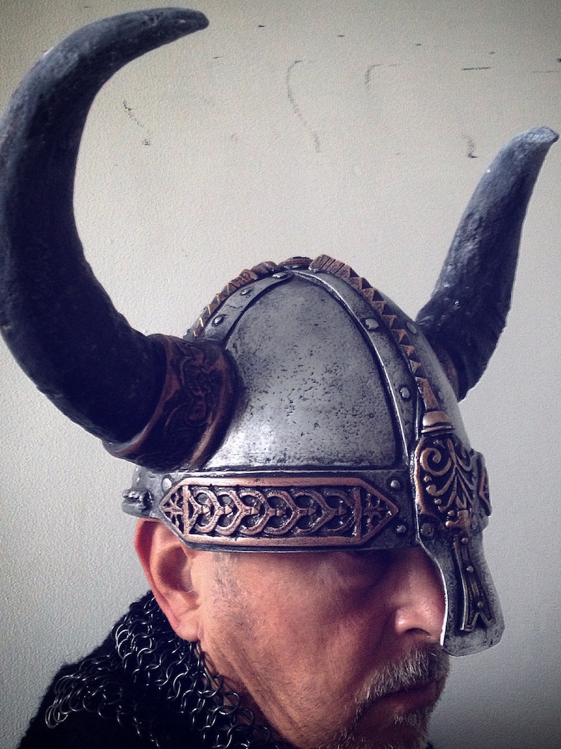 May include: A silver and copper Viking helmet with horns, worn by a person with a beard. The helmet has intricate details and a braided band.