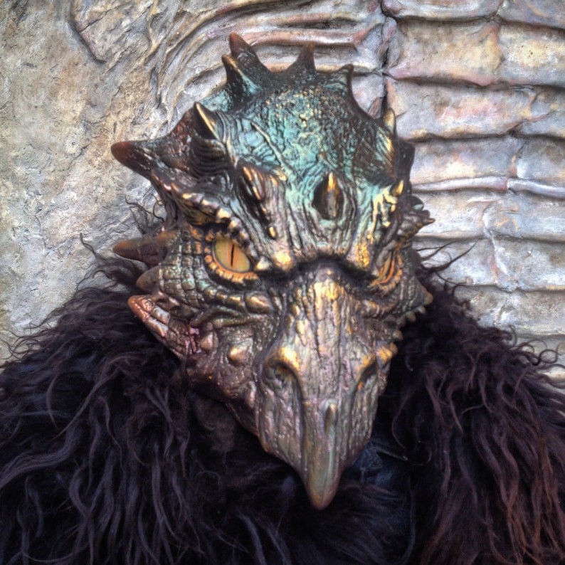 K&ouml;nnte beinhalten: Eine detaillierte, realistische Drachenmaske mit einer strukturierten, schuppigen Oberfl&auml;che. Die Maske ist in Gr&uuml;n-, Gold- und Braunt&ouml;nen bemalt, mit einem gro&szlig;en, goldenen Schnabel und gelben Augen.