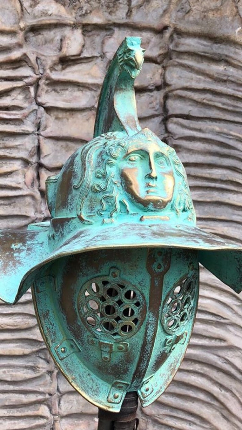 May include: A green and gold gladiator helmet with a face sculpted on the front. The helmet has a decorative crest on top and a metal grill over the face.