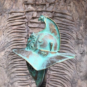 May include: A green and brown gladiator helmet with a plume on top. The helmet is on a stand in front of a textured background.