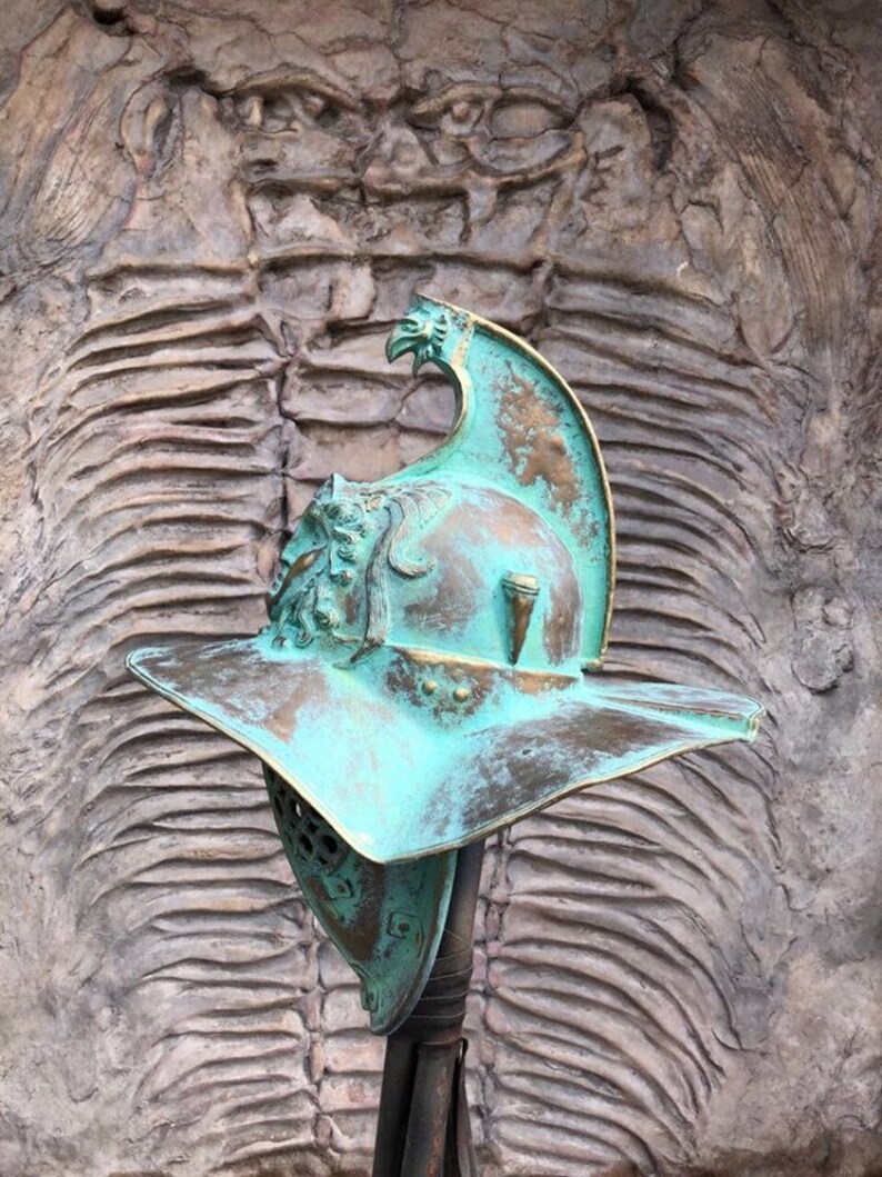 May include: A green and brown gladiator helmet with a plume on top. The helmet is on a stand in front of a textured background.
