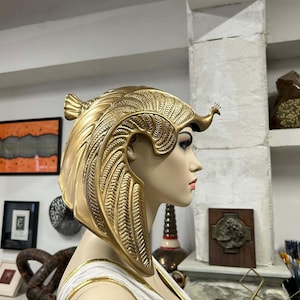 Peut inclure: Une coiffe dorée présentant des textures complexes en forme de plumes et une petite figurine d'oiseau sur le front, portée par un mannequin. La coiffe a une structure incurvée en forme d'aile qui descend le long de la tête et du cou.