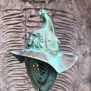 May include: A green metal gladiator helmet with a face mask and a crest on top. The helmet is on a stand against a textured background.