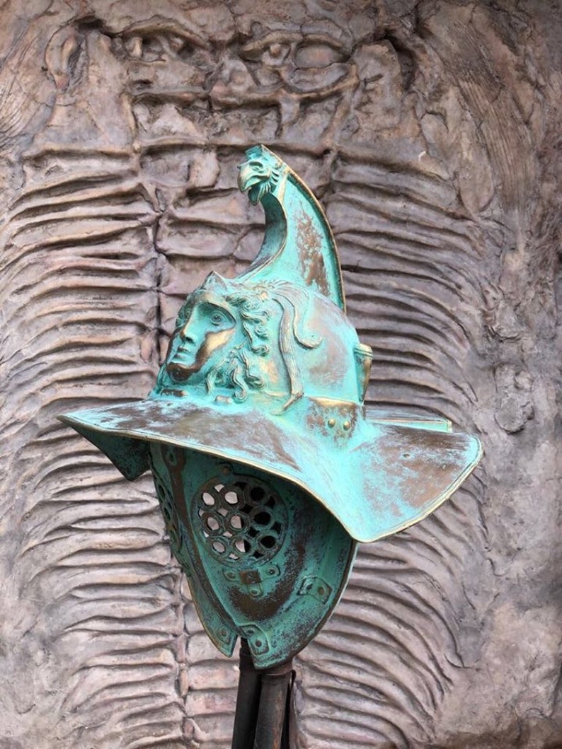 May include: A green metal gladiator helmet with a face mask and a crest on top. The helmet is on a stand against a textured background.