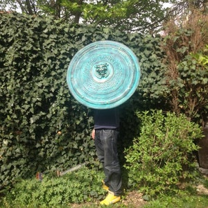 May include: A large, round, teal-colored shield with intricate designs and a central embossed face. The shield is held by a person wearing blue jeans and yellow shoes.