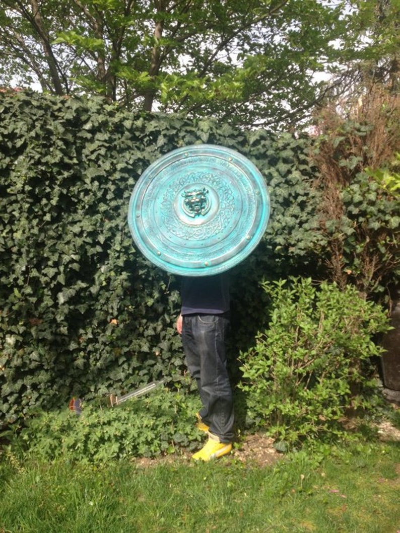 May include: A large, round, teal-colored shield with intricate designs and a central embossed face. The shield is held by a person wearing blue jeans and yellow shoes.