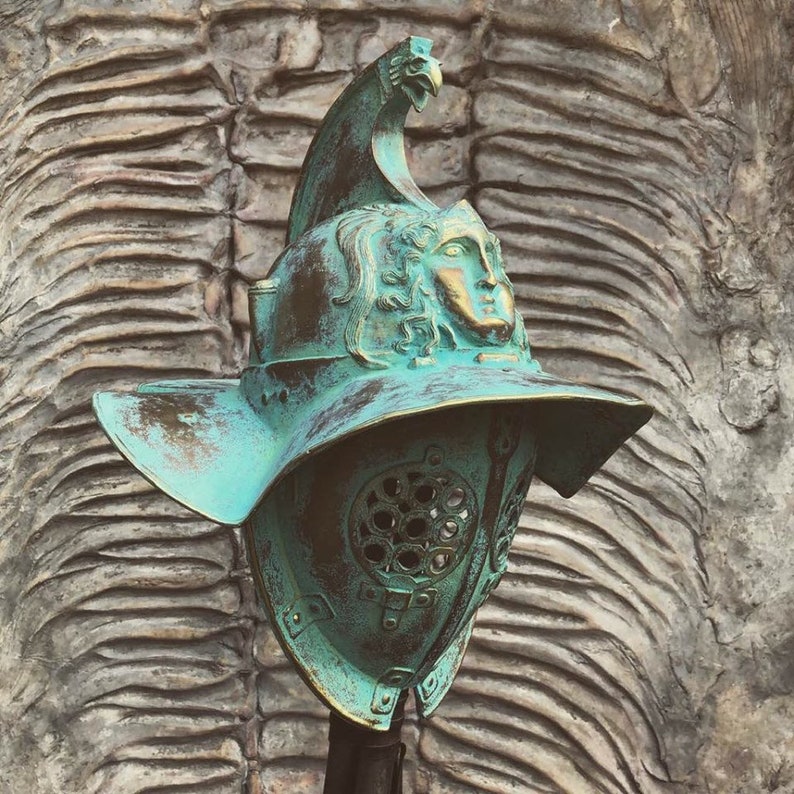 May include: A green and brown gladiator helmet with a face mask and a crest on top. The helmet is made of metal and has a distressed finish.