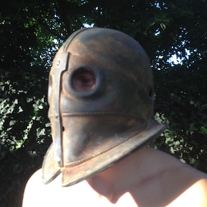 Puede incluir: Un casco de cuero marr&oacute;n con una banda de metal y dos agujeros para los ojos. El casco lo lleva una persona con los hombros descubiertos.
