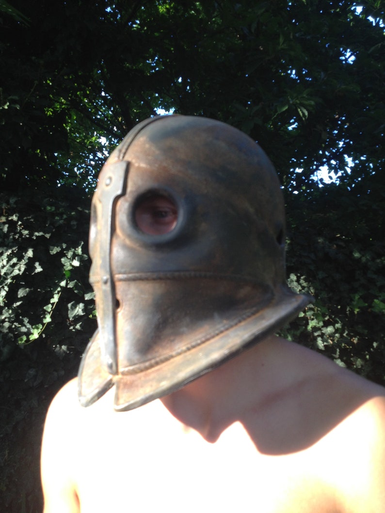 Puede incluir: Un casco de cuero marr&oacute;n con una banda de metal y dos agujeros para los ojos. El casco lo lleva una persona con los hombros descubiertos.