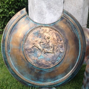 May include: A large, round, bronze-colored shield with a raised design of a mythical creature. The shield has a blue-green patina and a decorative border.