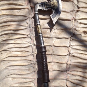 May include: A silver and black axe with a leather wrapped handle. The axe head has a decorative design with gold accents.